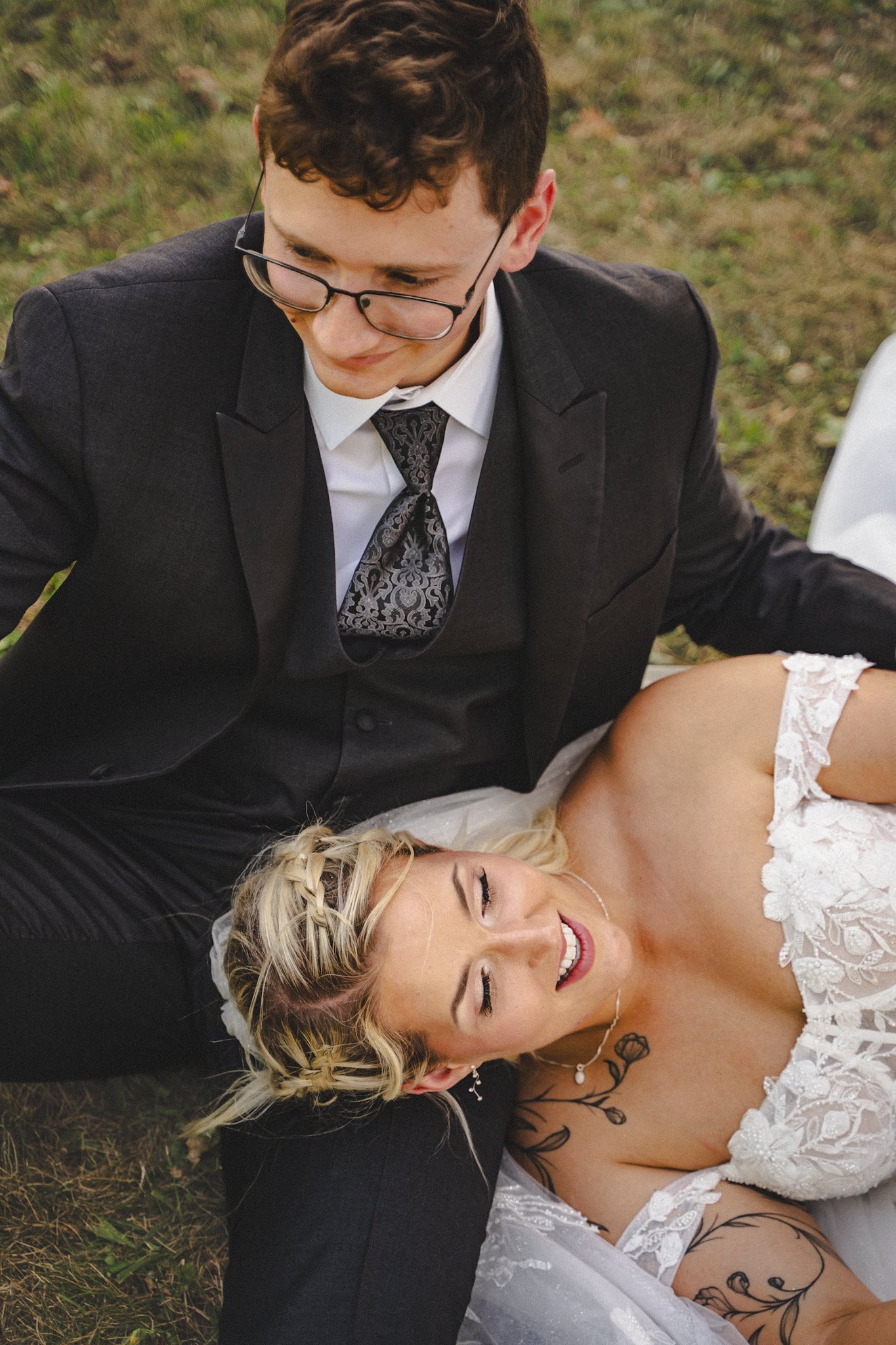A newlywed couple lying on the grass during their wedding photoshoot. The groom is dressed in a black suit with a patterned tie and glasses, and the bride is in a white lace wedding gown with tattoos visible on her arm. They are smiling and looking a