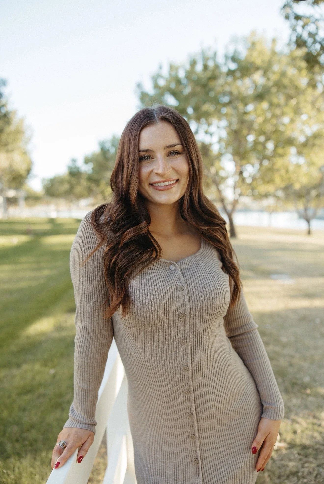 A woman with brown hair, blue eyes, and light skin, smiling, wearing a sleeveless black turtleneck top and beige pants, sitting on a cream-colored sofa in a well-lit room.