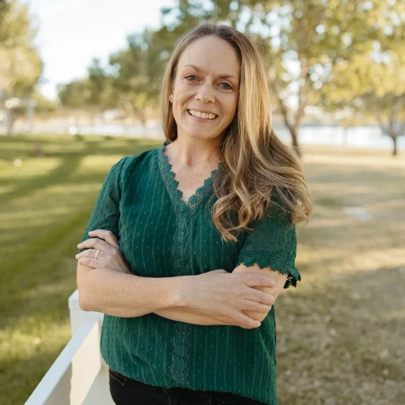 A woman with long, wavy blonde hair outdoors, smiling with her arms crossed, wearing a green top with lace details, with a park and trees in the background.