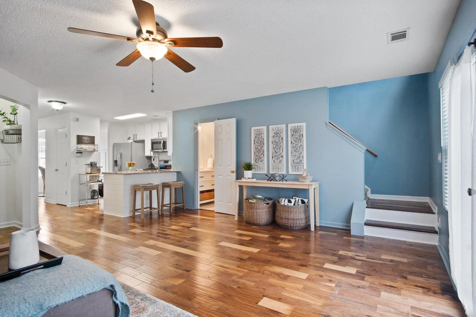 Interior view of a living room with a blue accent wall, staircase, and a kitchen with white cabinets and a granite countertop, hardwood flooring, ceiling fan, and natural lighting.