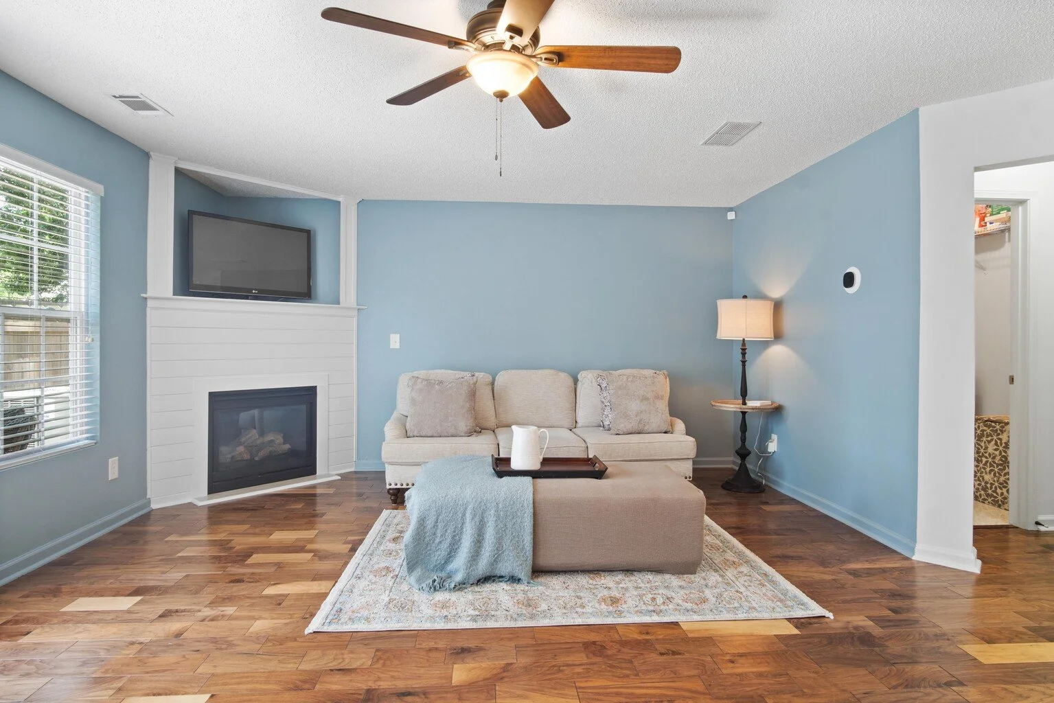 Living room with a light beige sofa, a small ottoman with a light blue throw blanket, a tray with a white pitcher, a floor lamp, a wall-mounted TV, and a fireplace on a blue wall. Hardwood flooring and a window with blinds are also visible.