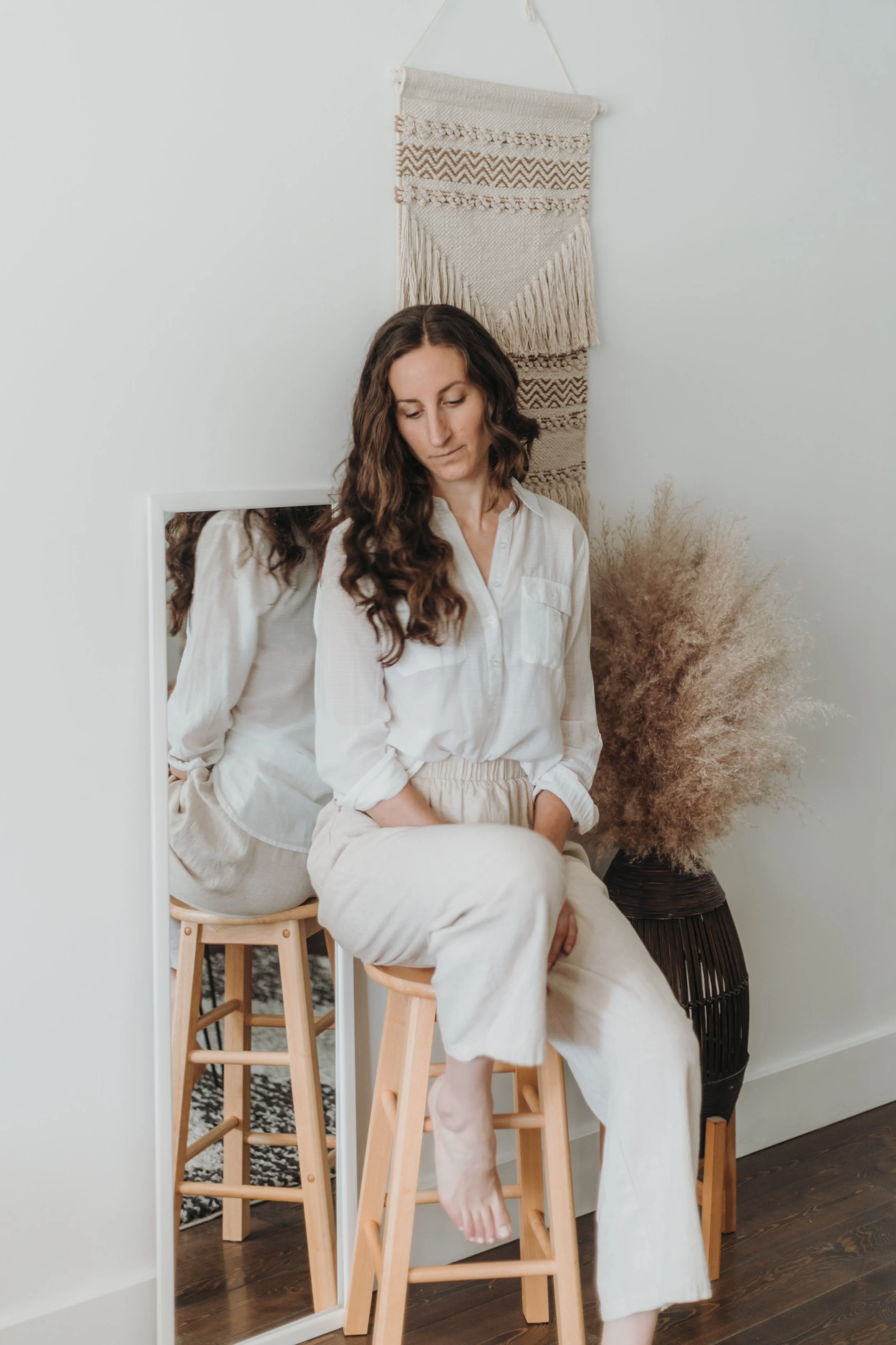 The author sits on a wooden stool wearing a white blouse and beige linen pants with a white wall background and dry grasses surrounding her. She looks down with a reflective stance. Women's coach, self-care, wellbeing, women's wellness.