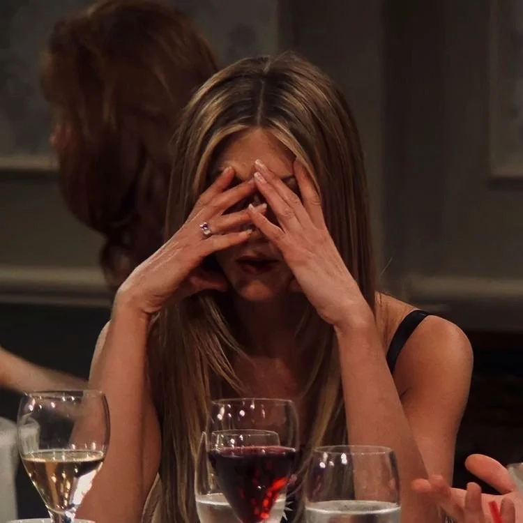 A woman with long hair sitting at a table with drinks, covering her face with her hands.