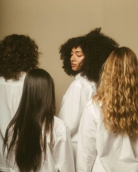 Four people with diverse hairstyles wearing white shirts, standing together. One person faces forward with curly hair, while the others have their backs turned. The background is neutral.
