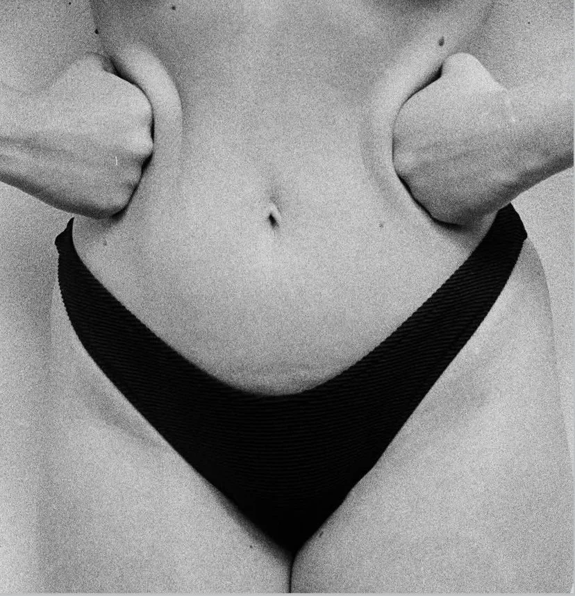 Close-up of a woman's torso showing her navel and hips, with her hands clenched into fists on her sides, wearing black underwear, in black and white photography.