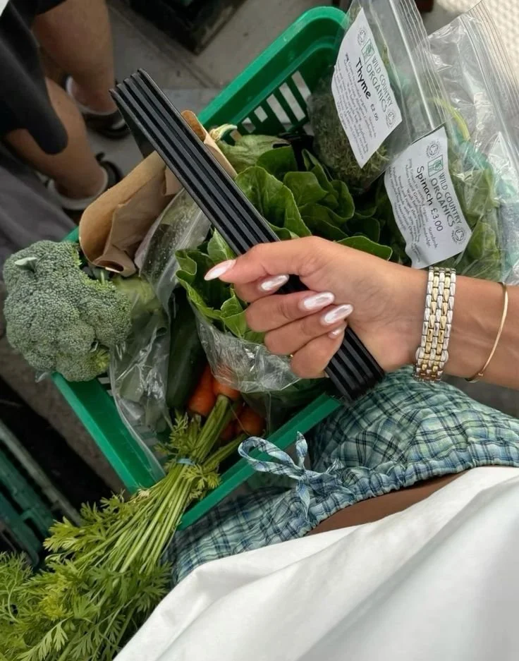 A person holding a green shopping basket filled with fresh vegetables, including broccoli, spinach, carrots, and a bundle of herbs, at a grocery store.