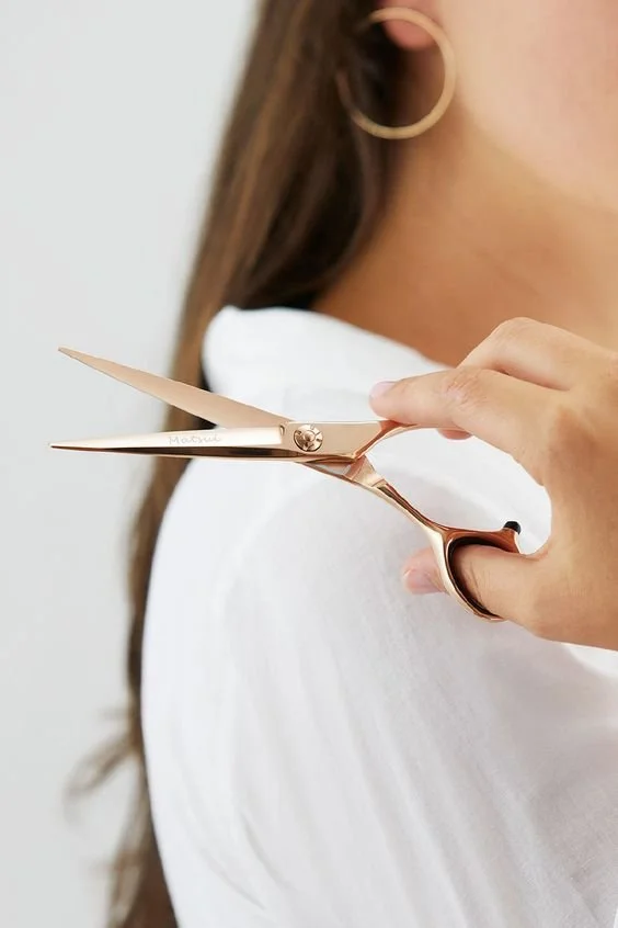 Person holding gold scissors wearing white shirt and hoop earrings.