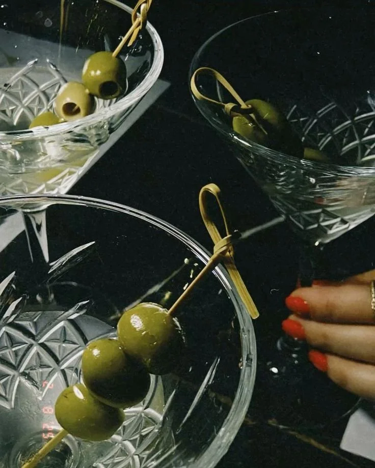 Close-up of three glass bowls with green olives each on a skewer, some with pimento filling, on a dark surface, with a person's hand with red nail polish visible.
