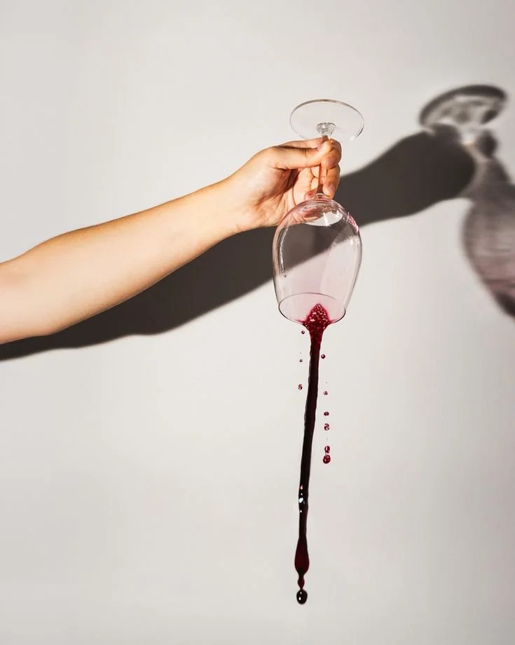 A hand holding an upside-down wine glass with red wine spilling out, creating a shadow on the wall.