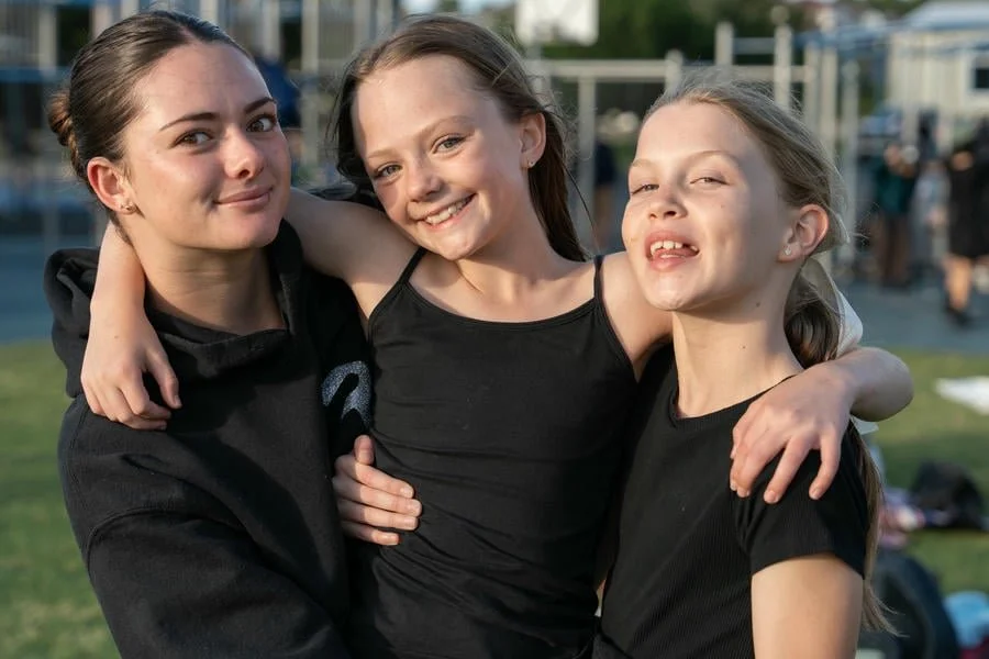 Three young girls smiling and hugging each other outdoors at a park or event on a sunny day.