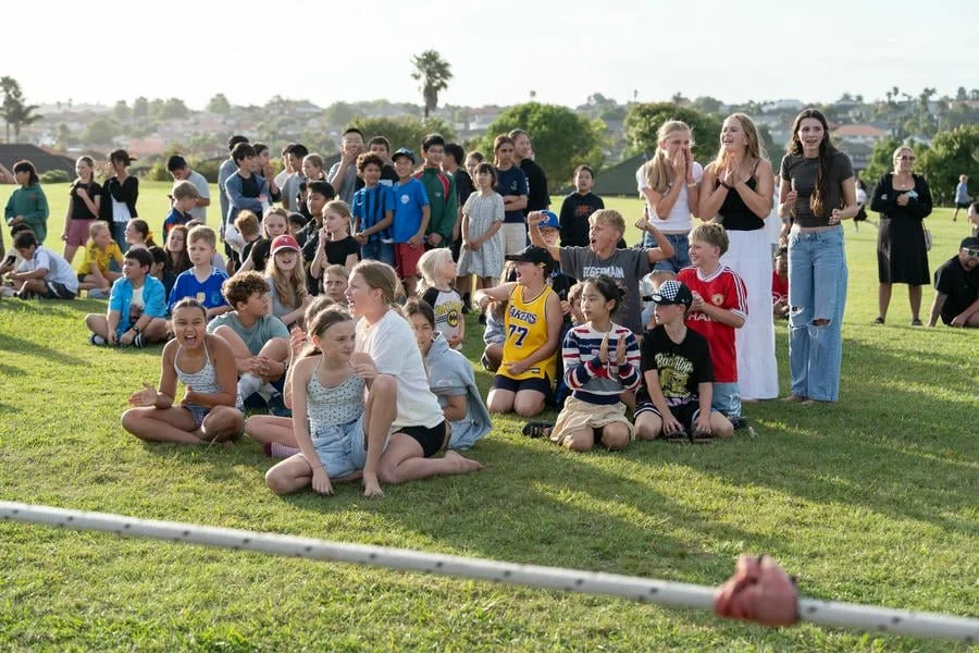 Group of children and adults sitting and standing on a grassy field, watching an outdoor event under a clear sky with some trees and houses in the background.