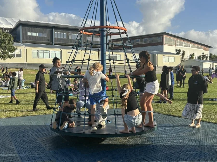 Children playing on a carousel at an outdoor park on a sunny day with an audience and modern buildings in the background.