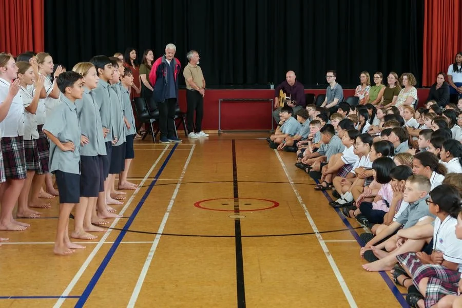 School assembly with students sitting on the floor and standing, in a gymnasium with a stage.