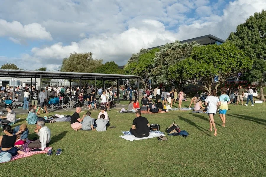 People gathered outdoors at a park or event on a sunny day, some sitting on the grass and others standing or walking near trees and a covered pavilion.