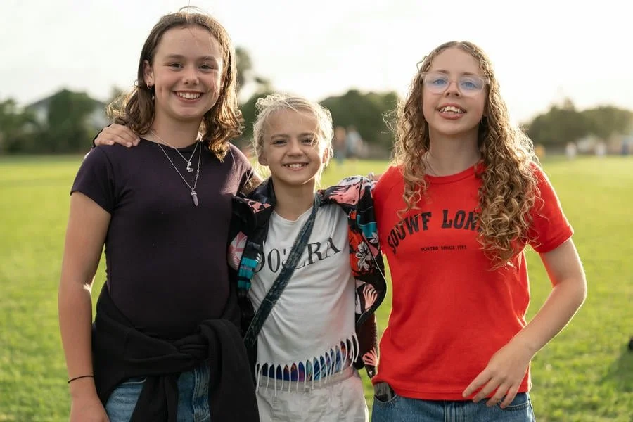 Three girls standing on a grassy field, smiling at the camera, with trees and a sunny sky in the background.
