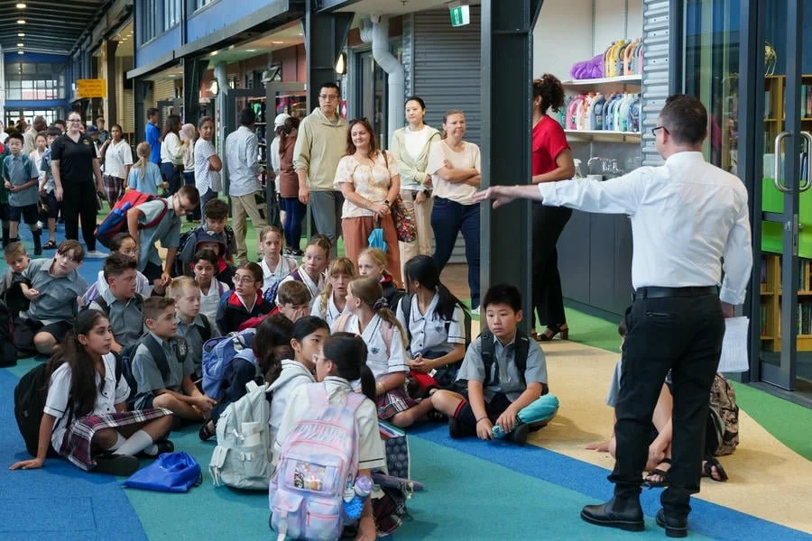 A large group of students sitting on the floor inside a shopping mall, listening to a man in a white shirt and black pants speaking. Several adults are standing around, observing the scene.
