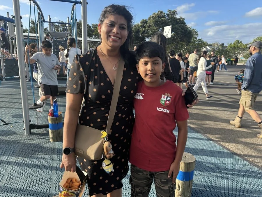 A woman and a boy standing together at an outdoor playground, smiling. The woman is wearing a black dress with pink polka dots, and the boy is wearing a red sports jersey. There are many people in the background, some children playing and adults walk