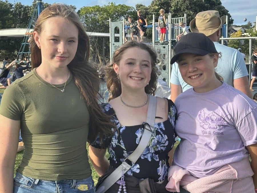 Three girls standing close together outdoors, smiling, with a playground in the background and other children playing.