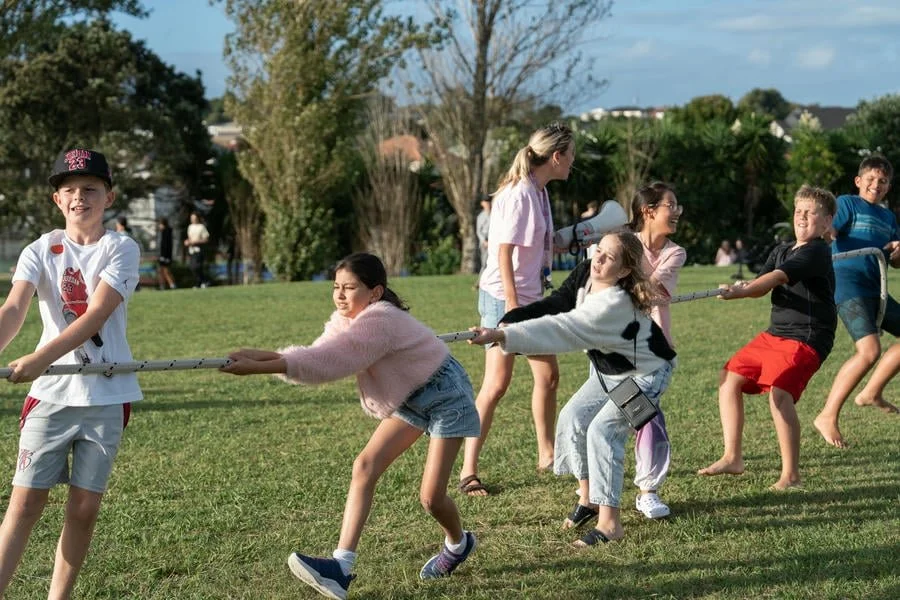 Children and a woman playing tug of war on a grassy field in a park.