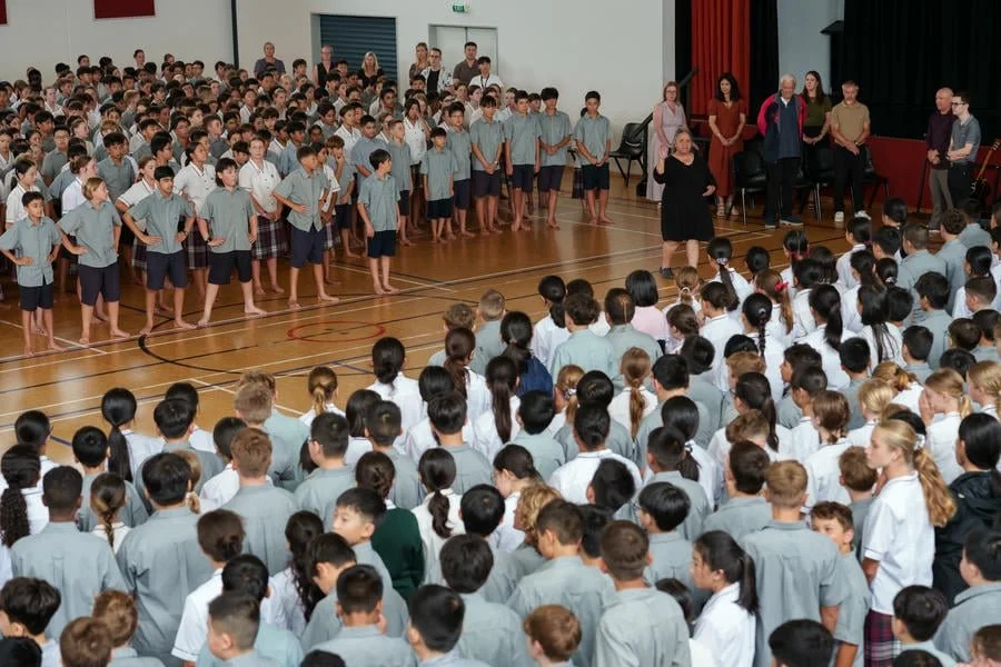 Large group of students facing a stage in a school gymnasium, with a few teachers or staff members standing in the background, during a school assembly or performance.