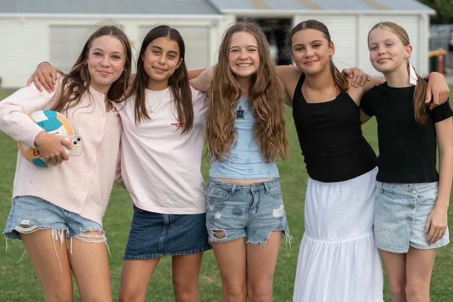 Five girls standing on grass with arms around each other, smiling at the camera, outdoors during daytime.