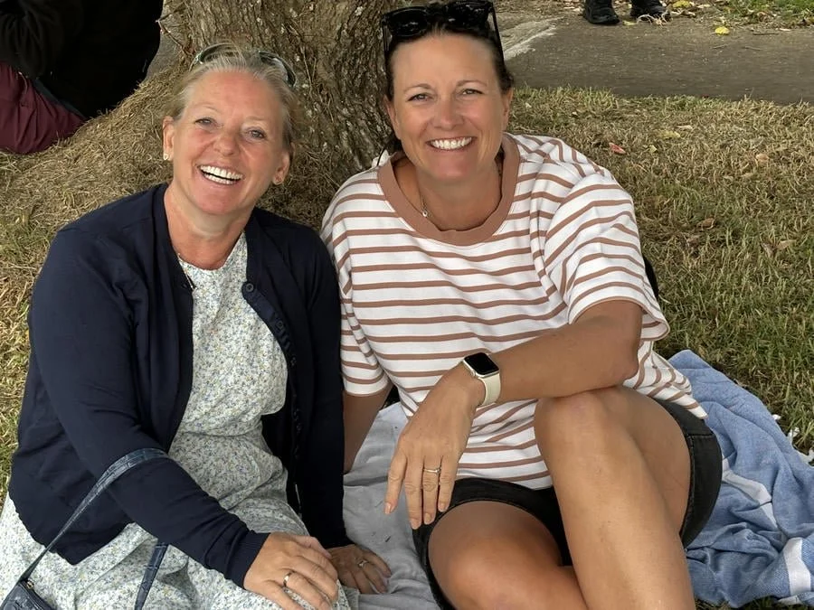Two women sitting under a tree, smiling at the camera, one wearing a dark jacket and dress, the other in a striped shirt and shorts.