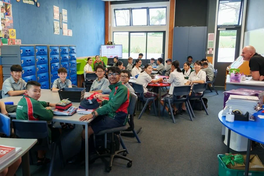 A classroom full of middle school students sitting at tables, some working on laptops, with posters on the blue walls and lockers in the background, during daytime.