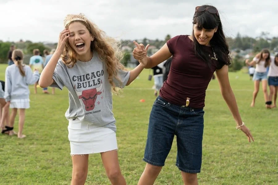 Two girls dancing and laughing on a grassy field with other people in the background.