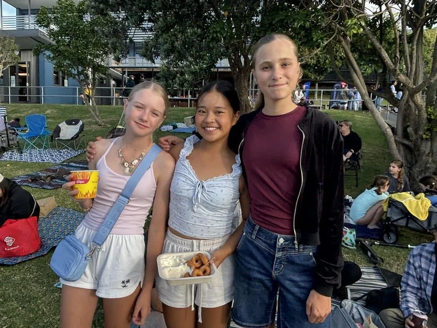 Three young girls standing together in a park during a gathering, with picnic blankets and people in the background.