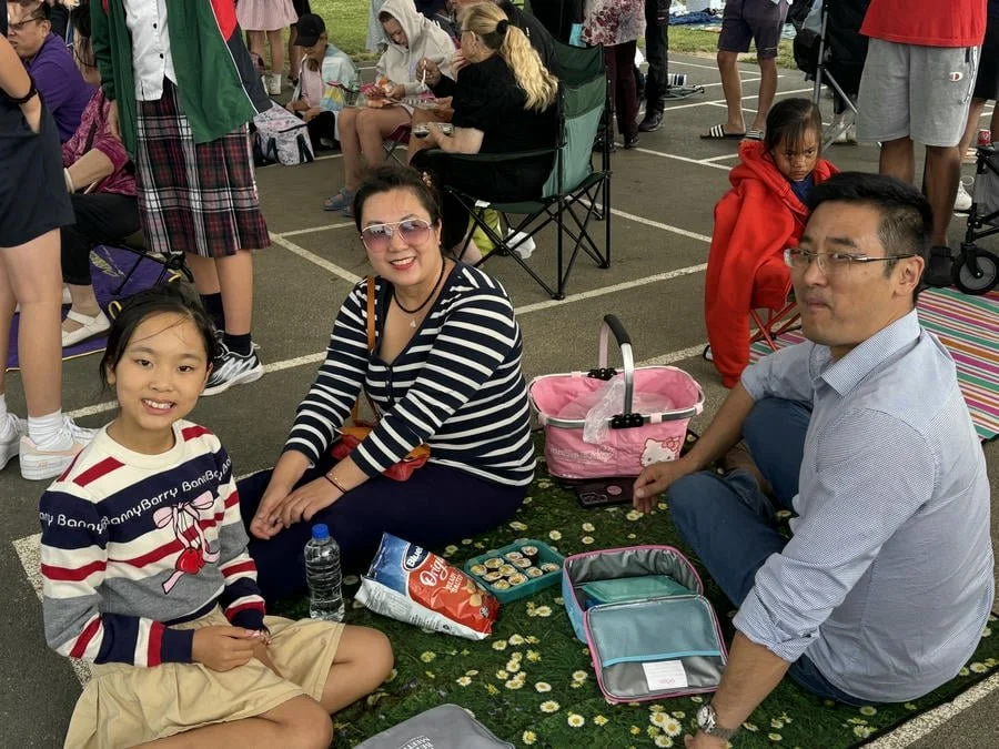 A family of three sitting on a picnic blanket at an outdoor event, with people and chairs in the background. The young girl is wearing a striped sweater, the woman is wearing sunglasses and a striped shirt, and the man is dressed in a light shirt and
