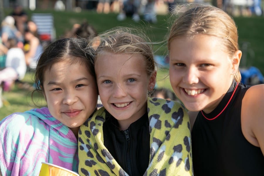 Three young girls smiling outdoors on a sunny day, with a crowd and green grass in the background.