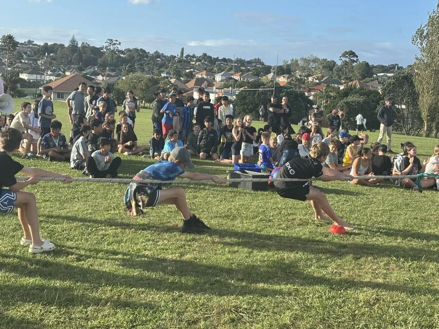 Children participating in a tug-of-war game on a grassy field with spectators in the background and a neighborhood view. The children are pulling on a rope, leaning back, with some adults watching.