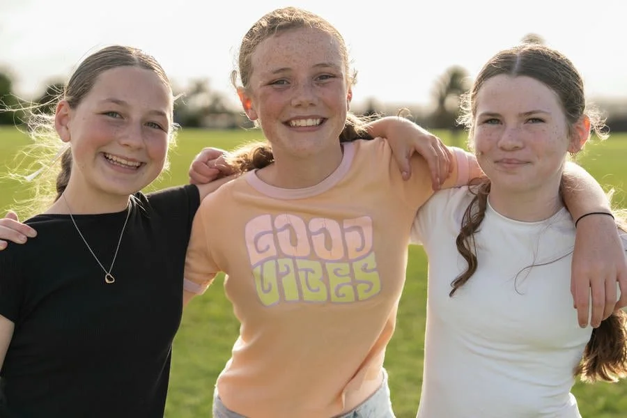Three young girls standing outdoors on a sunny day with arms around each other, smiling.