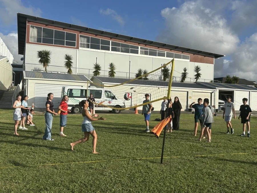 Children and teenagers playing volleyball on a grassy field outside a modern building with palm trees, vehicles, and a partly cloudy sky in the background.