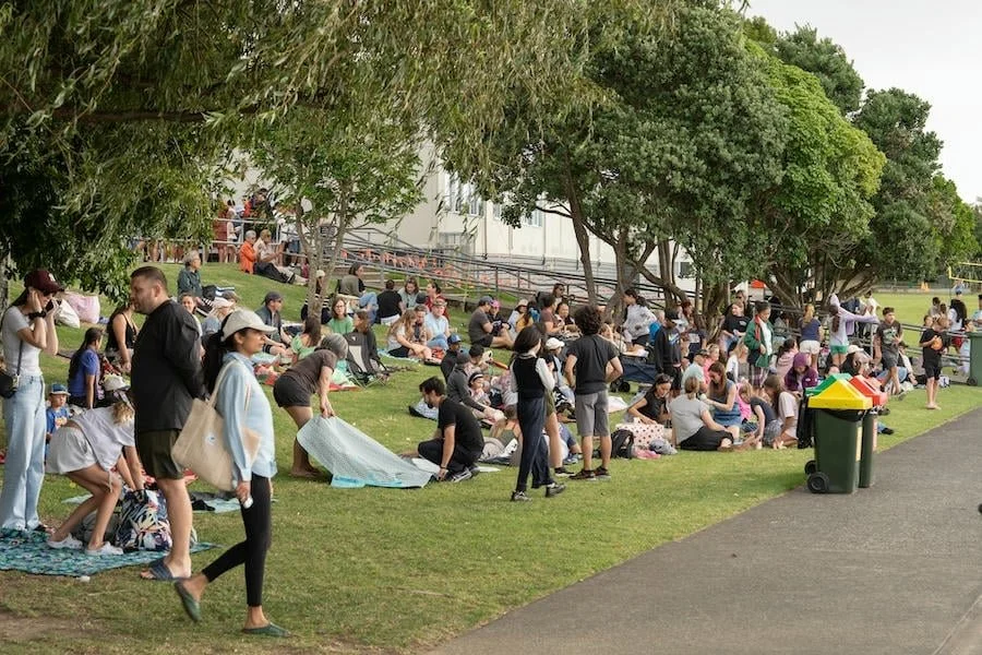 A large group of people sitting on the grass and on blankets under trees in a park, engaging in an outdoor event or gathering.