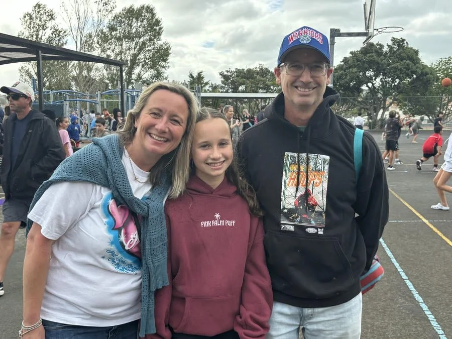 Group of three people smiling at a school basketball court during a game or event, with children playing in the background.
