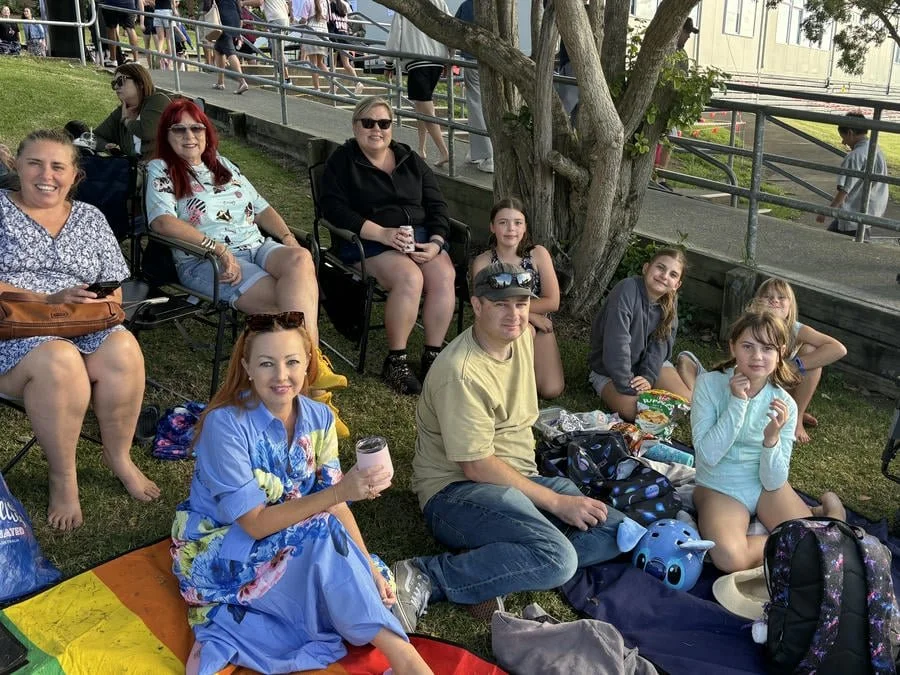 Group of adults and children relaxing near a tree outdoors, some sitting on lawn chairs and blankets, with snacks and drinks.