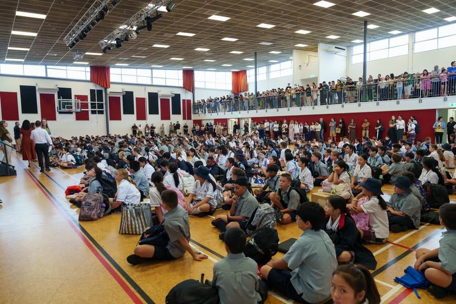 Students sitting cross-legged on the gymnasium floor, some raising their hands, attending a school assembly with many spectators watching from an upstairs balcony.