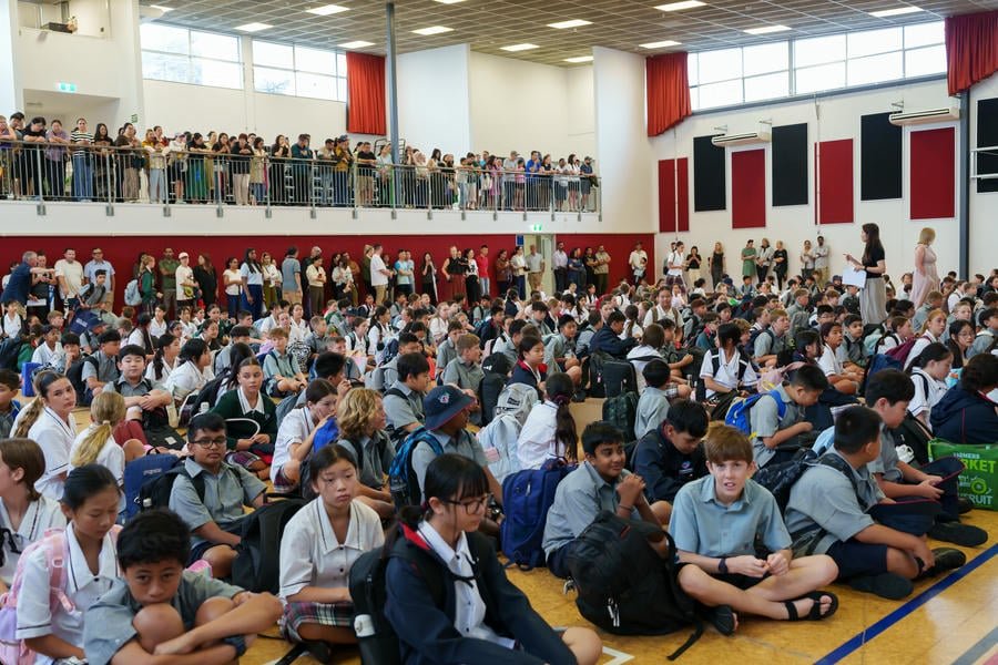A large group of students seated on the gymnasium floor, with more standing along the balcony railing, in a school assembly or event setting.