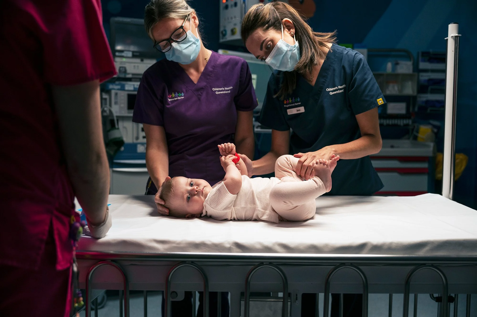A baby lying on an examination table while medical staff, including two women wearing scrubs and masks, examine the baby in a hospital setting.