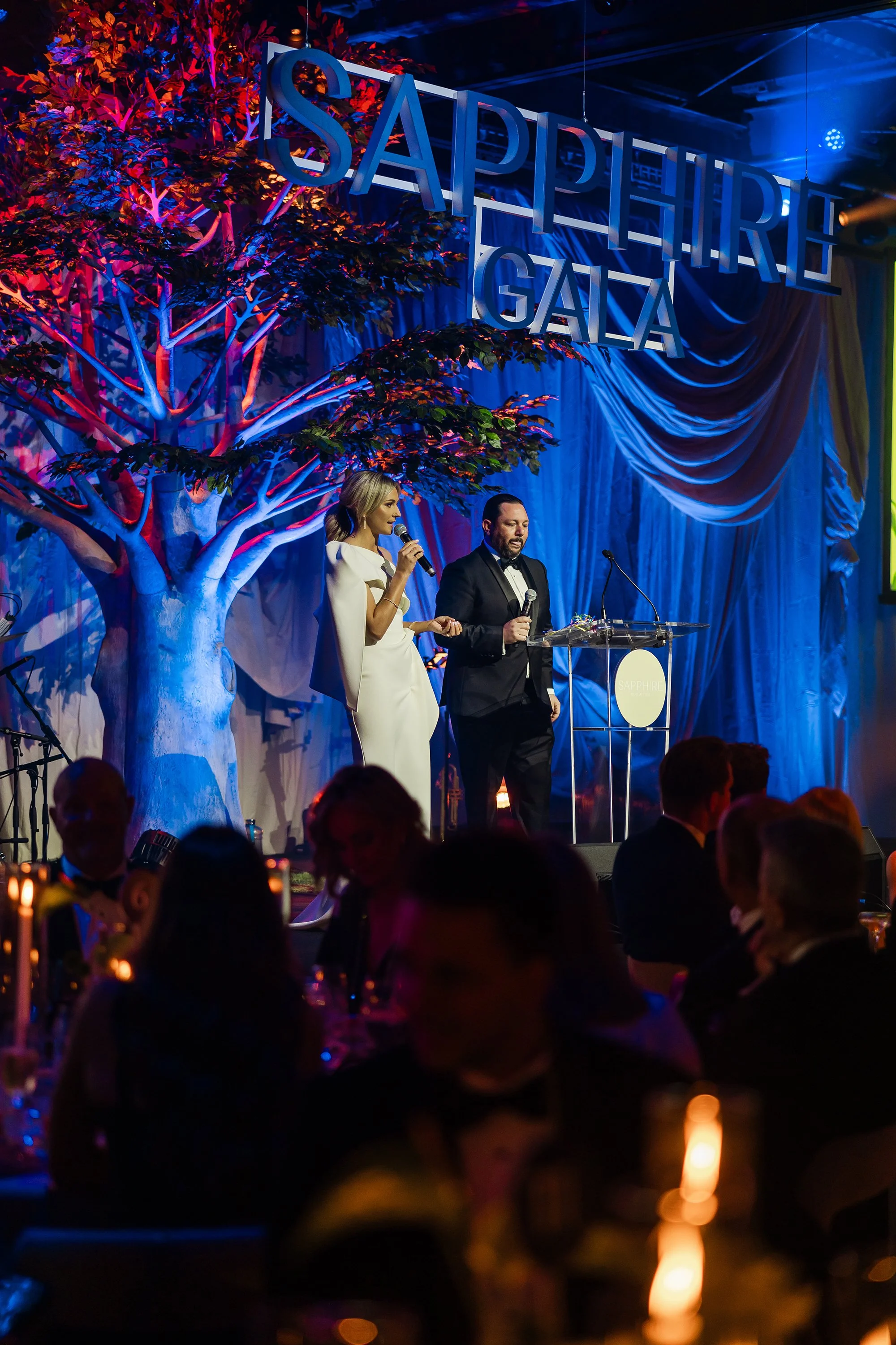 Two hosts, a woman in a white gown and a man in a black tuxedo, speaking at a formal event on stage with a large sign reading 'Sapphire Gala' and a decorative tree with colorful lighting in the background.