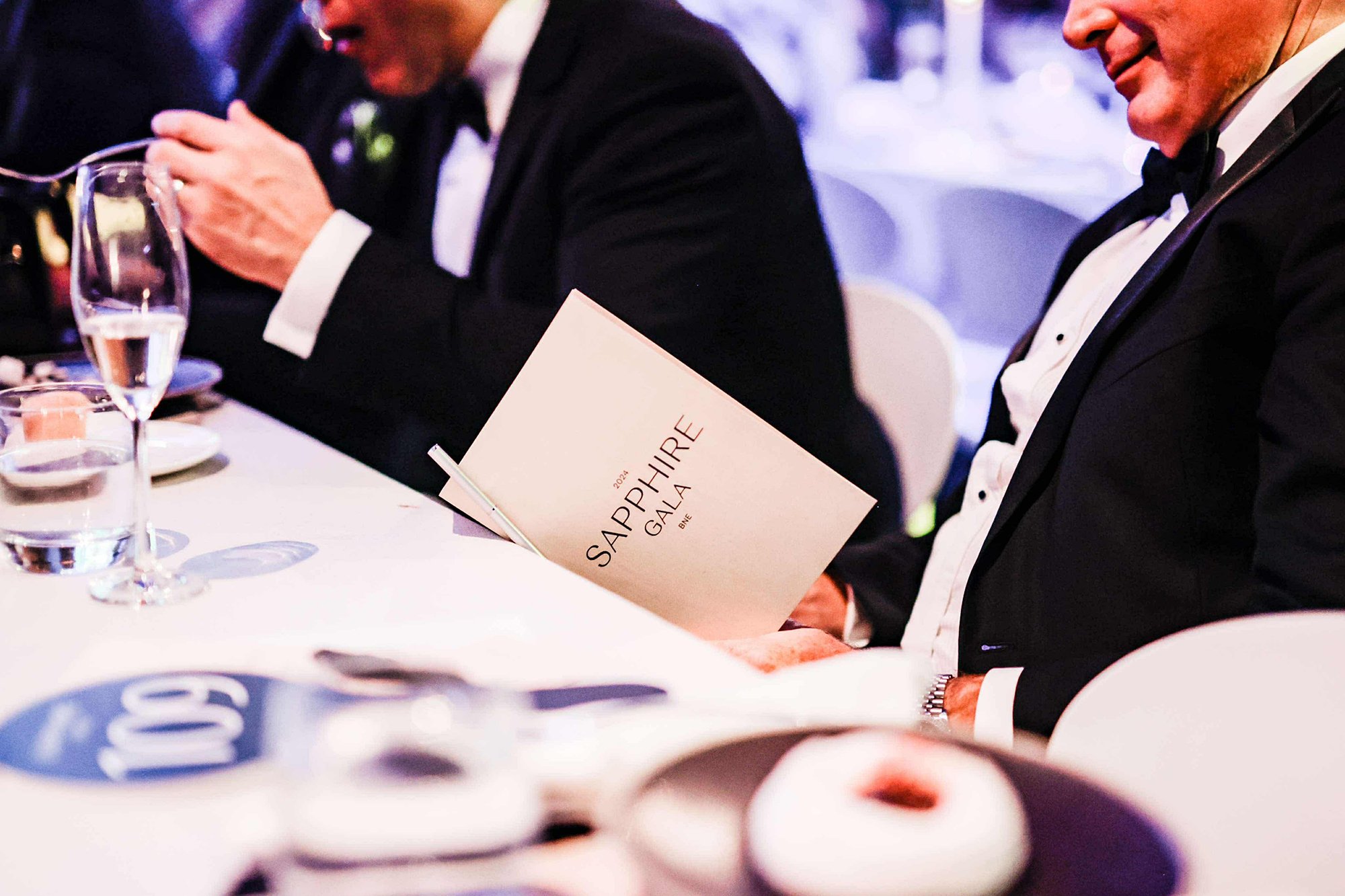 Men in tuxedos sitting at a formal banquet table with drinks and pastries, one man reading a program titled 'Sapphire Gala 2024'.