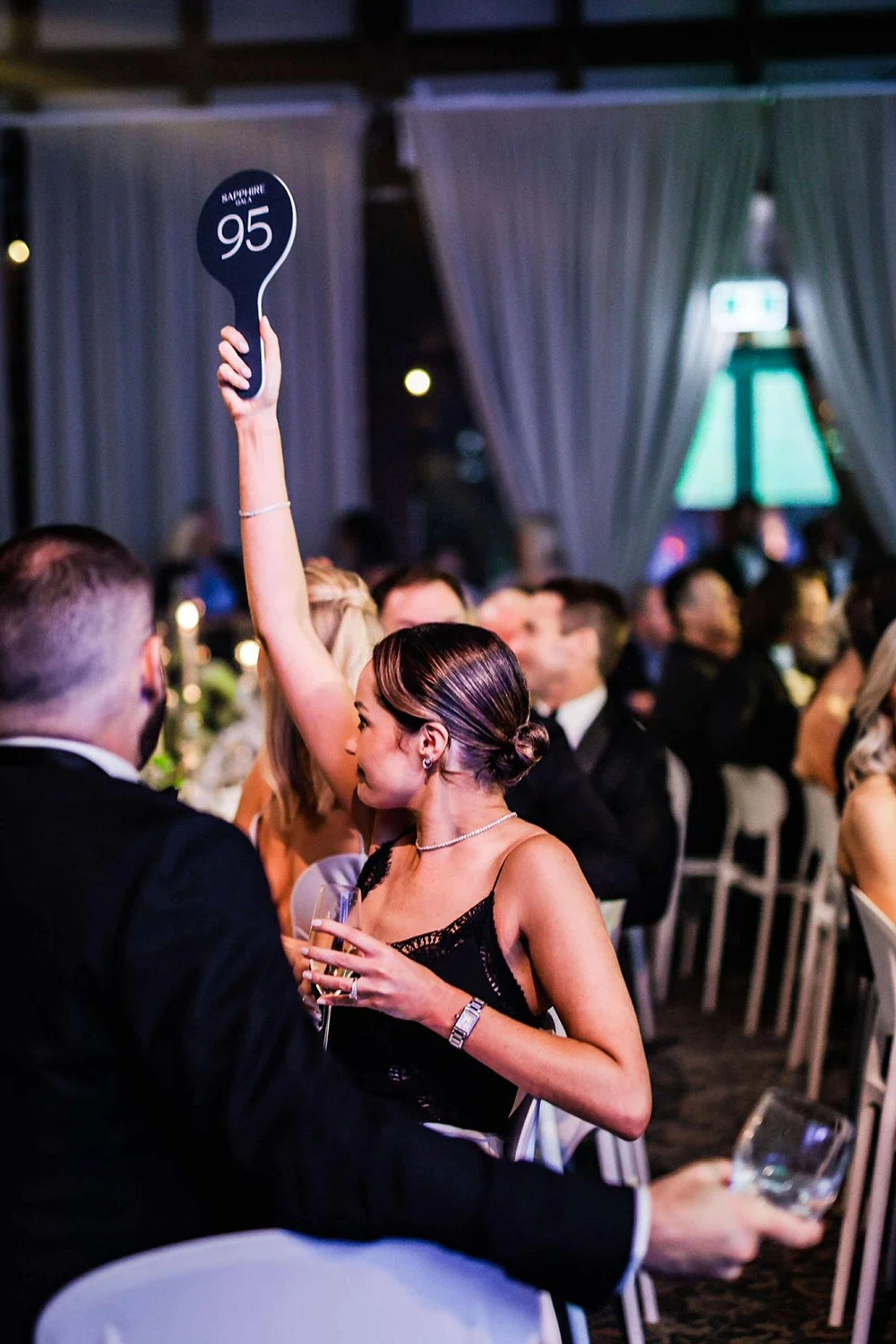 Woman holding up paddle with '95' written on it at a seated auction or event, surrounded by other attendees in formal attire, dimly lit indoor setting.