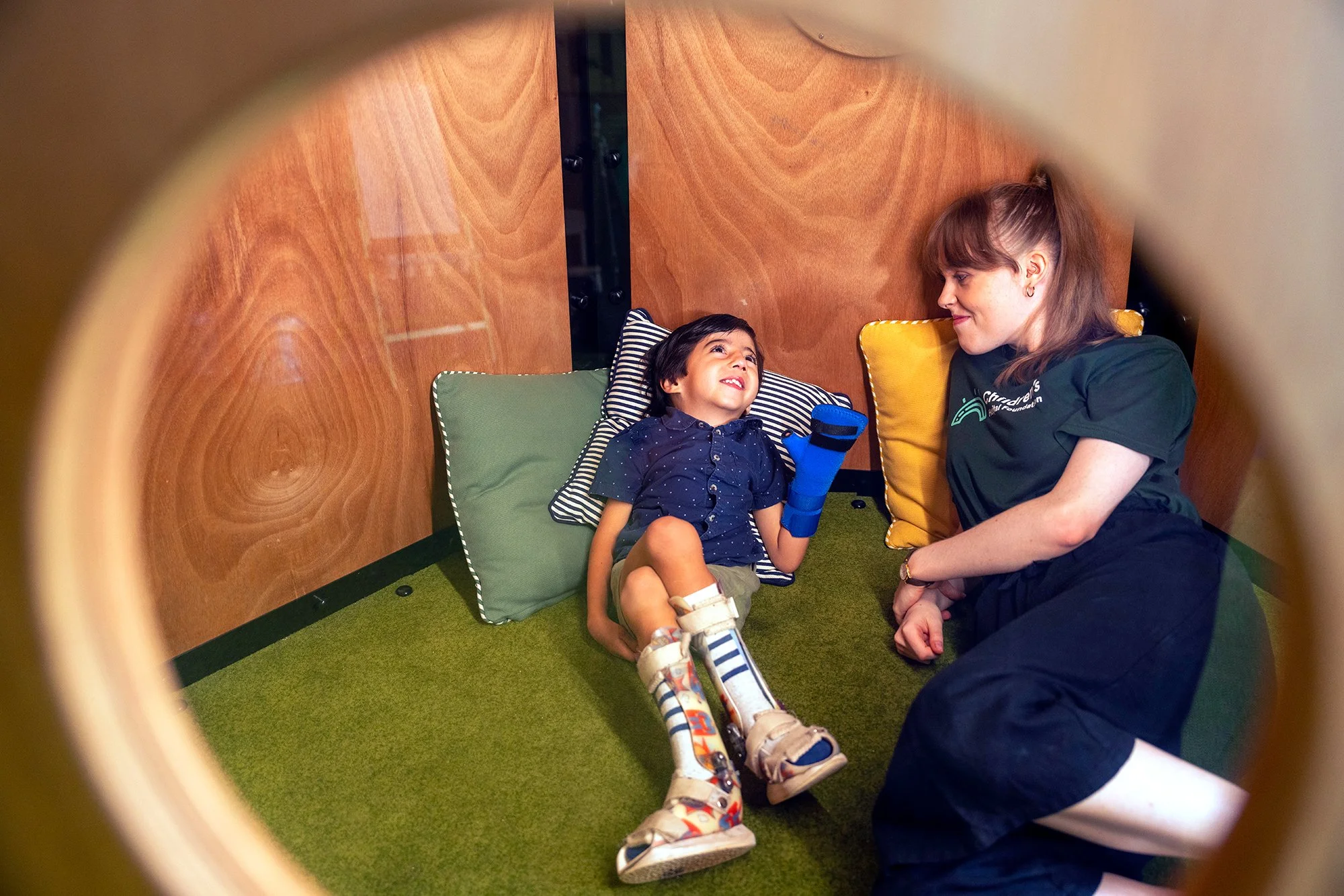 A young boy with a cast on his arm is lying on the floor, talking to a woman, possibly a caregiver, in a therapy or medical setting. The boy is smiling and wearing colorful socks and sneakers, while the woman is sitting beside him, smiling back.