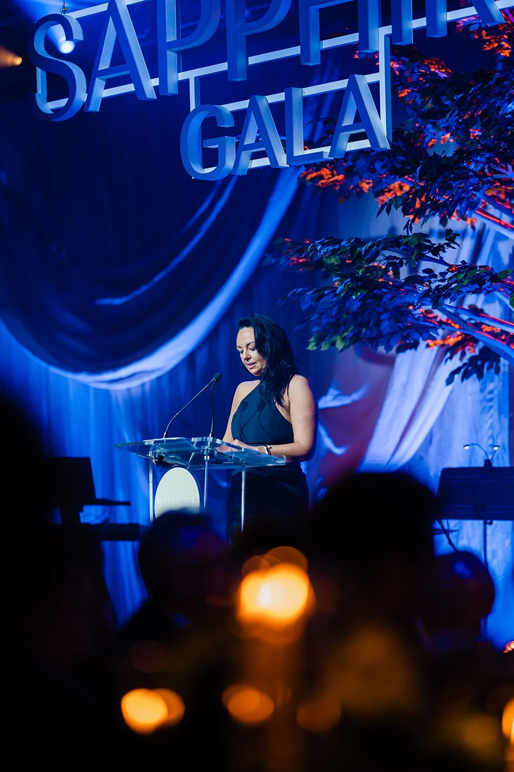 A woman in a black dress giving a speech at a podium during a Sapphire Gala event. Large 'Sapphire Gala' sign hangs above, with stage decorated with drapery and a tree with orange and green leaves illuminated by stage lights.
