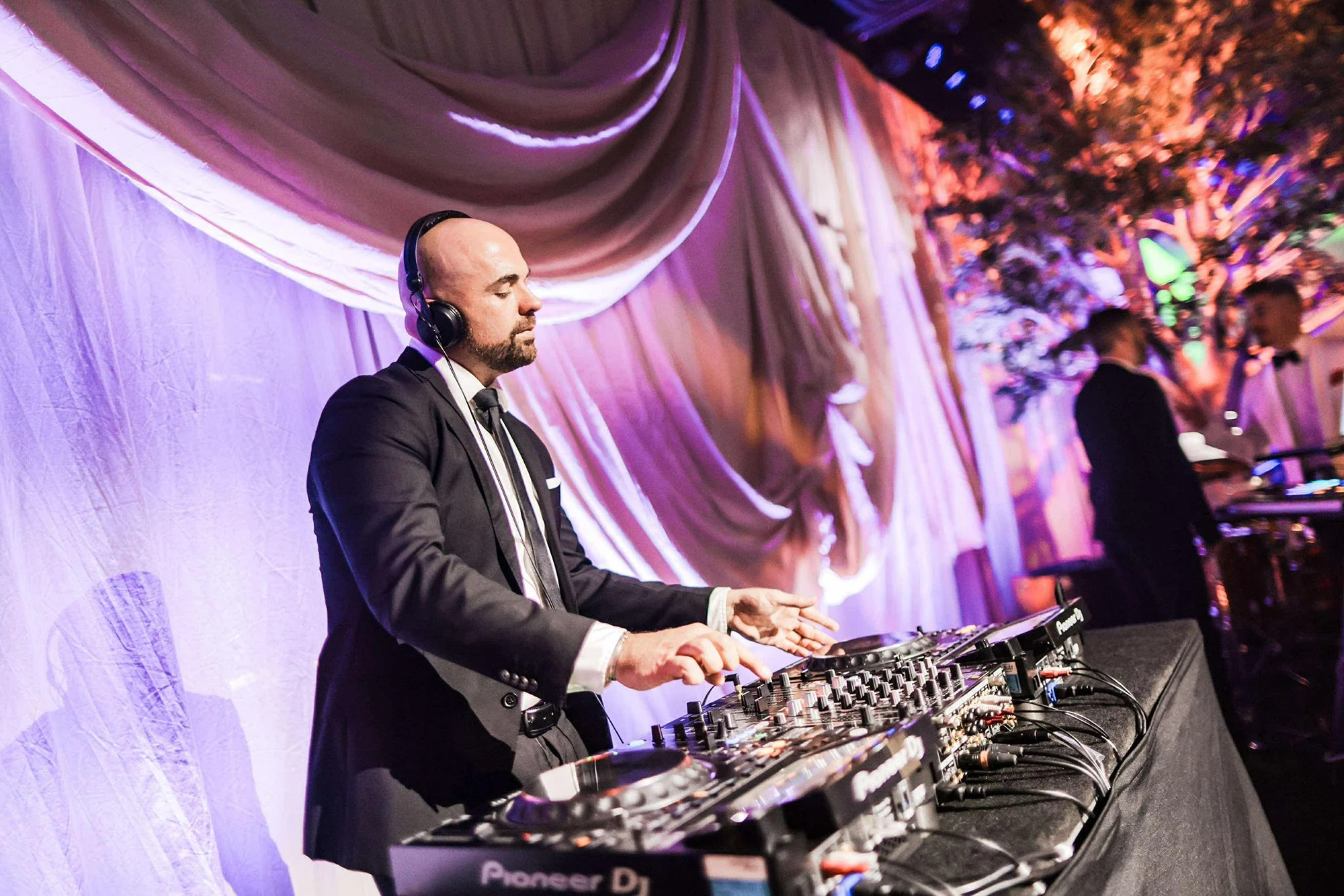 A DJ in a black suit and white shirt DJing at an event, wearing headphones and standing behind a Pioneer DJ mixer. The background features draped fabric and purple and pink lighting with blurred people.