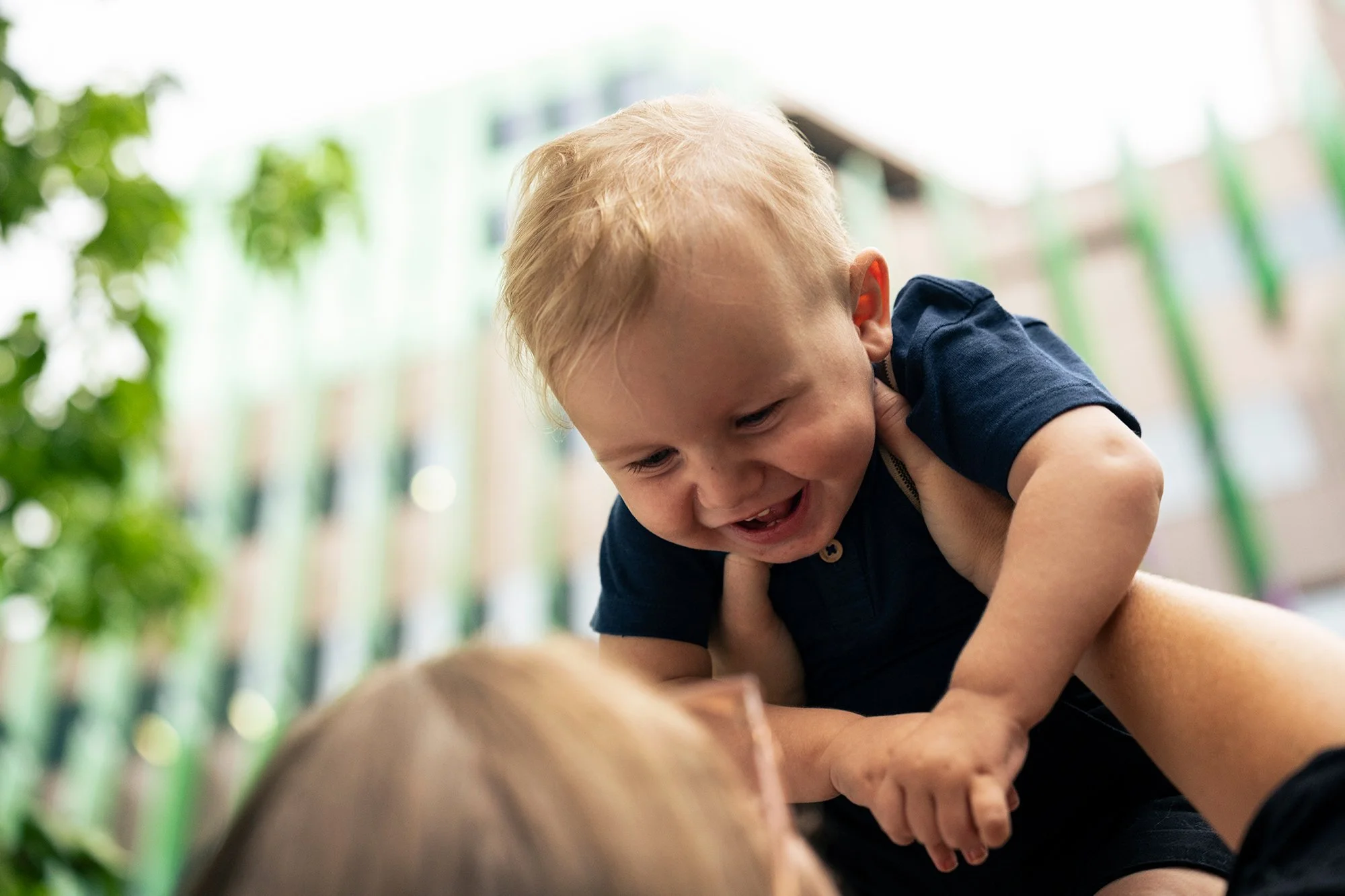 A young boy with blonde hair playing a game of tickle fight with an adult, smiling and showing joy, outdoors with green trees and a building in the background.