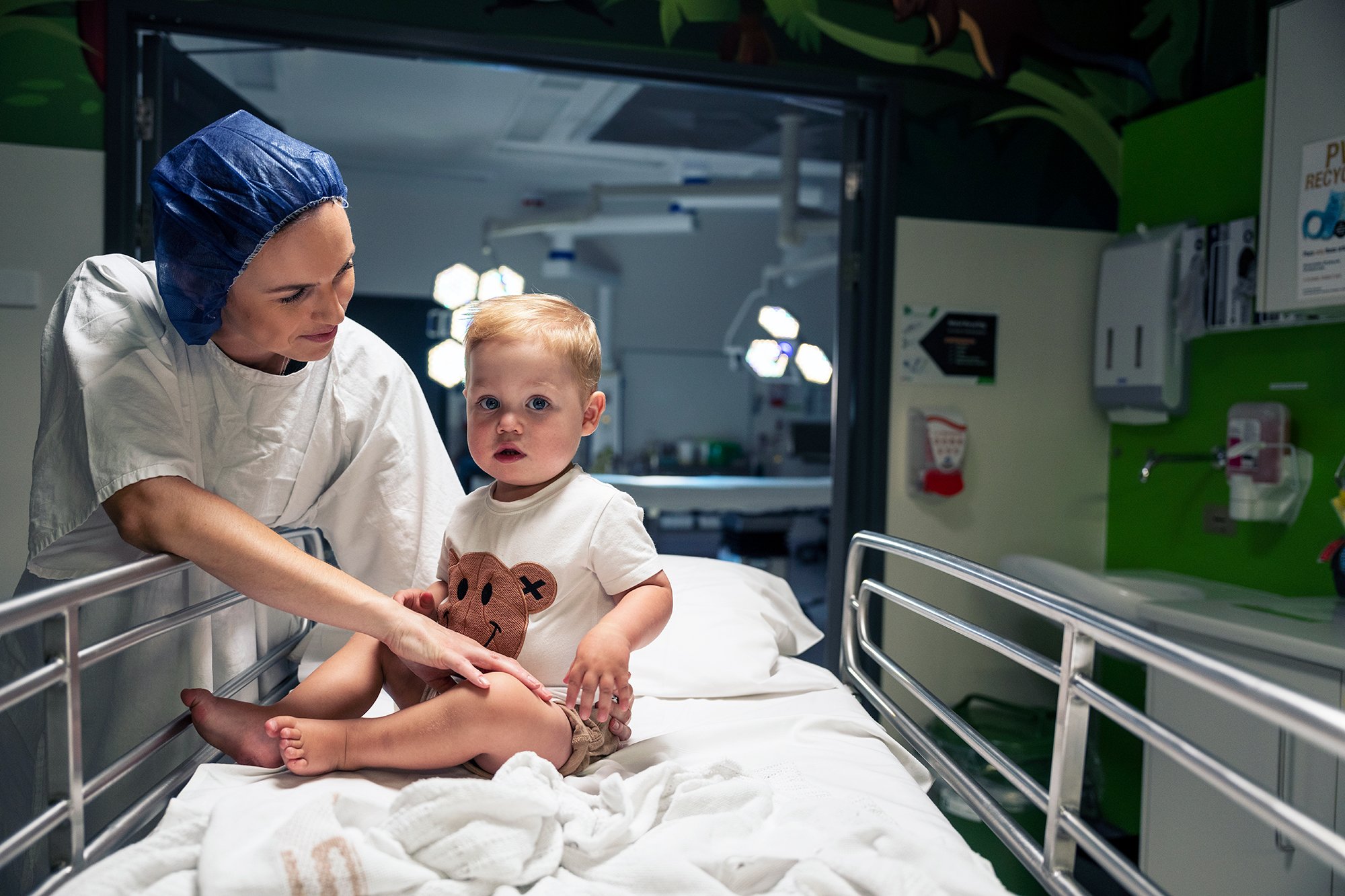 A nurse or doctor in medical scrubs and a blue hair cap examining a young girl sitting on a hospital bed. The girl has blonde hair, blue eyes, and is wearing a T-shirt with a bear face graphic. The hospital room has medical equipment and bright overhead lights.