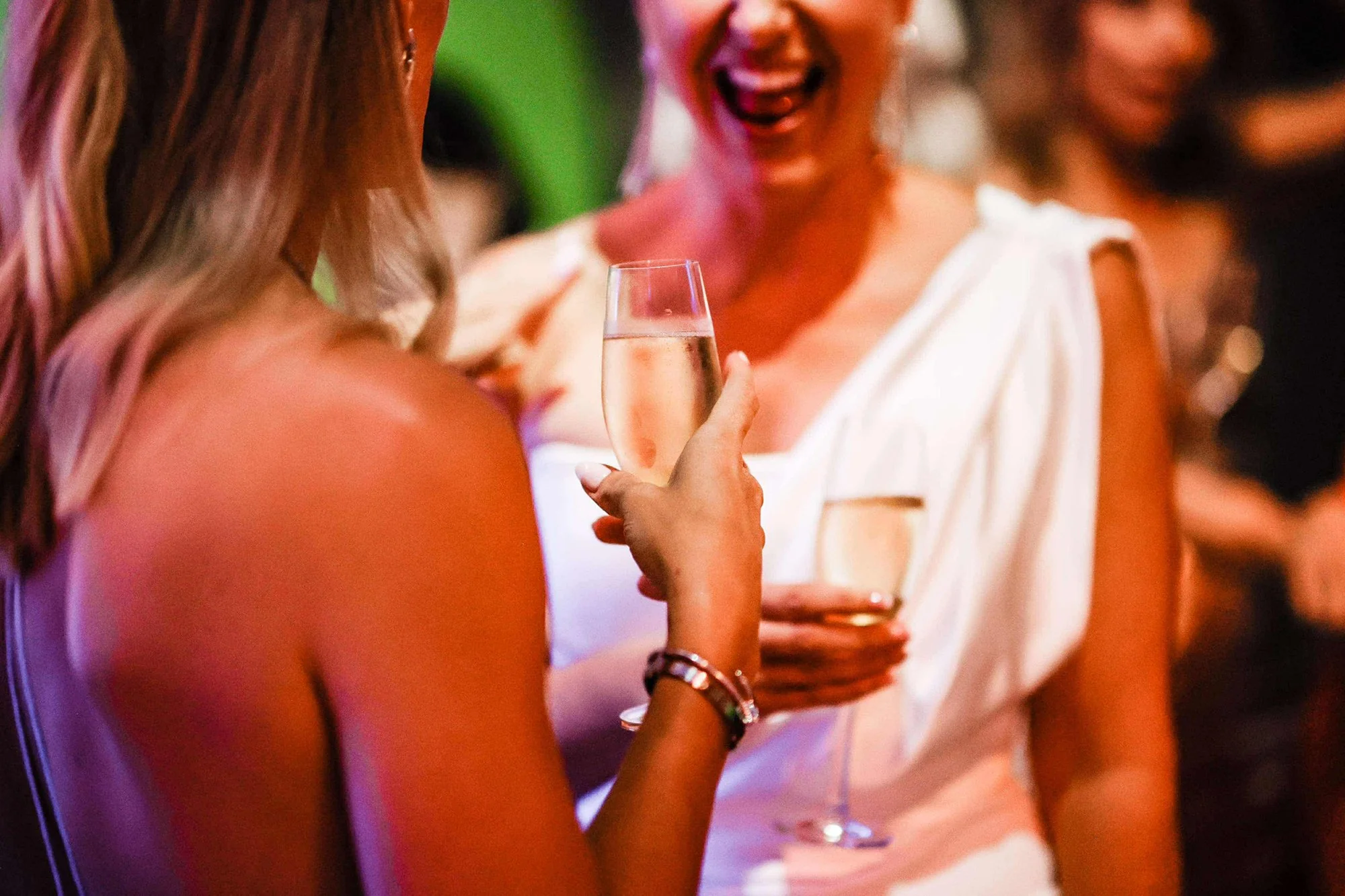 Two women enjoying a social event, one holding a glass of champagne or sparkling wine, engaging in conversation and smiling.