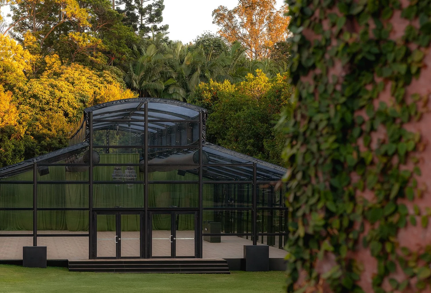 A glass greenhouse or event space with black framing, surrounded by lush greenery and trees with fall foliage.